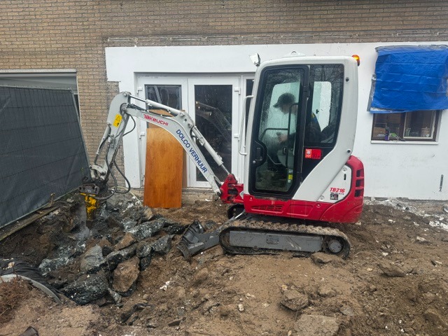Minigraver met sloophamer breekt bestrating en fundering open naast een woning